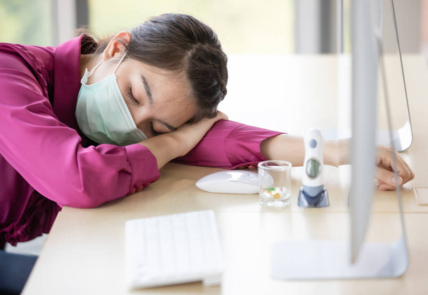 Θολή φωτογραφία της εργαζόμενης γυναίκας που φοράει προστατευτική μάσκα υγιεινής και εργάζεται στο γραφείο και αισθάνεται άρρωστος πυρετός στη συνέχεια κουρασμένος και να πέσει κάτω και να κοιμηθεί στο τραπέζι. Threre είναι ιατρική και ψηφιακό θερμόμετρο στο γραφείο. - Φωτογραφία, εικόνα