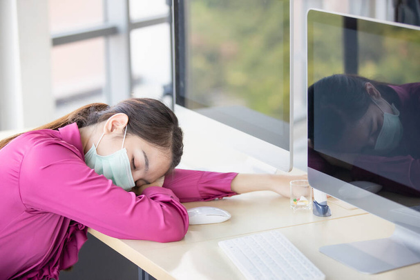 Rozmazane zdjęcie pracowniczki noszącej maskę higieniczną pracującej w biurze, źle się czującej, zmęczonej, upadającej i śpiącej na stole. Threre są medycyna i termometr cyfrowy na biurku. - Zdjęcie, obraz