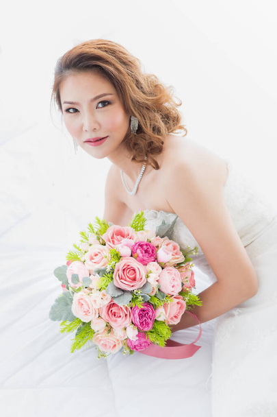 Ritratto primo piano della bella sposa asiatica seduta nello spogliatoio con un mazzo di fiori in grembo con sfondo di tende bianche
. - Foto, immagini