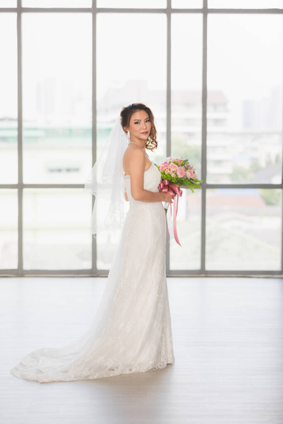 Вертикальный портрет крупным планом с красивой азиатской невестой, стоящей и держащей букет цветов в гримерке
. - Фото, изображение
