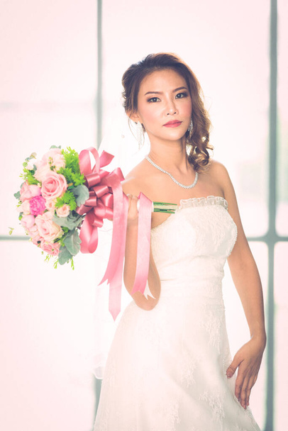 Крупный план портрет красивой азиатской невесты, стоящей и держащей букет цветов в гримерке
. - Фото, изображение
