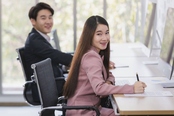 Asian businessman and businesswoman sitting, smiling to camera, woman holding pen, in office. - Photo, Image
