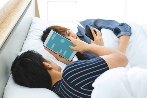 Adorável casal asiático deita-se na cama. Ele está a usar um tablet para conversar com ela. Há uma bolha de fala em branco no telemóvel dela. Eles não falam uns com os outros por via oral, mas usam tecnologia.
. - Foto, Imagem