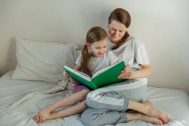 Happy young mother with daughter are reading a book in the light bedroom. They look at the book together. Family relationships and home leisure. Good mood. Free space. Horizontal photo. - Fotografie, Obrázek