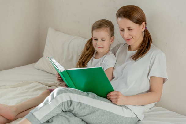 Happy young mother with daughter are reading a book in the light bedroom on a sunny morning. They look at the book together. Family relationships and home leisure. Good mood. Free space - Foto, Bild