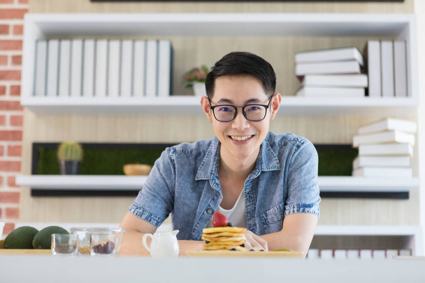 Detailní záběr portrét pohledný asijský muž se šťastnou tváří sedí v čisté kuchyni. Před ním je talíř s palačinkou, který ukazuje, jak důležité je jeho jídlo.. - Fotografie, Obrázek
