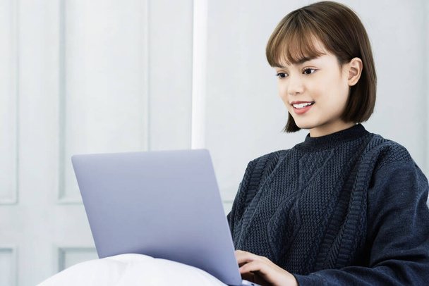 美しいアジアの女性は、家のベッドの上に座ってノートパソコンで作業します.ソーシャルアプリや仕事をチェック女性.ホームコンセプトから仕事. - 写真・画像