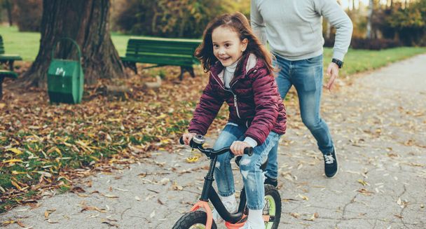Lovely girl riding the bike and trying to escape from her father running after her in an autumn day - Foto, Bild