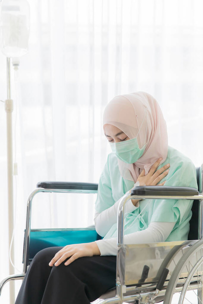 Femme musulmane portant un masque chirurgical d'hygiène de protection assis sur un fauteuil roulant pour le patient à l'hôpital, utiliser la prise des mains et toucher le cœur et le sentiment d'anxiété. Idée pour les entreprises de santé
. - Photo, image
