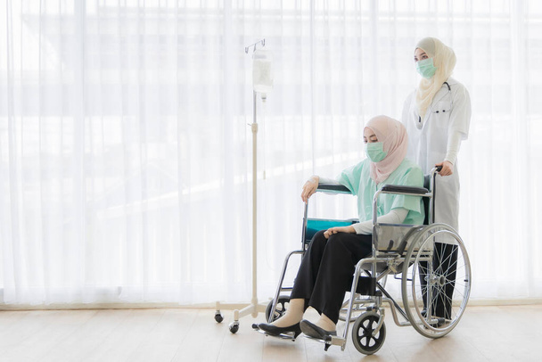 Médecin musulmane en robe hijab aide gentiment la patiente qui est assise en fauteuil roulant à l'hôpital. Ils portent un masque protecteur d'hygiène chirurgicale sur le visage pour les protéger des virus et des maladies
. - Photo, image