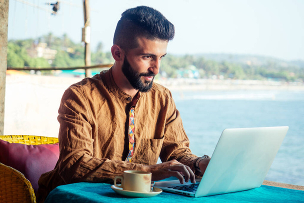Egyptian happy man with laptop computer drinking coffee or masala chai at beach shack in Goa - Foto, immagini