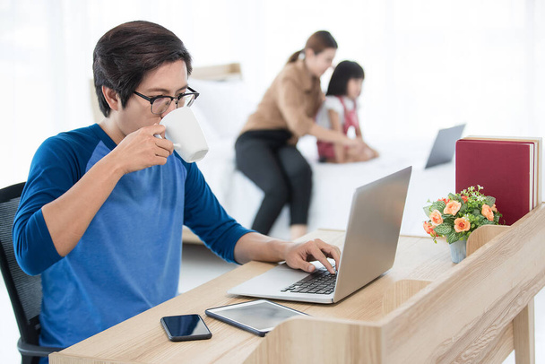 Azjatycki biznesmen pije kawę podczas pracy w domu z komputerem, tabletem i smartfonem, podczas gdy jego rodzina, żona i córka uczą się z laptopa w rozmytym tle. Koncepcja pracy w domu. - Zdjęcie, obraz