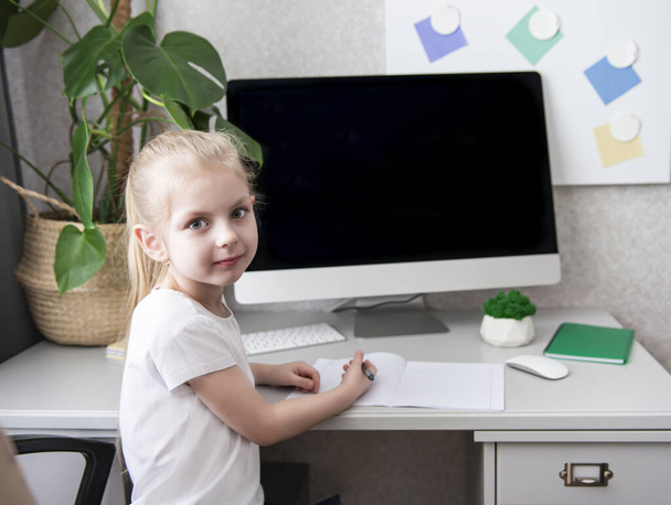 Залишайтеся вдома, щоб запобігти пандемії коронавірусу. Маленька дівчинка зі світлим волоссям сидить за столом і робить домашнє завдання
.  - Фото, зображення