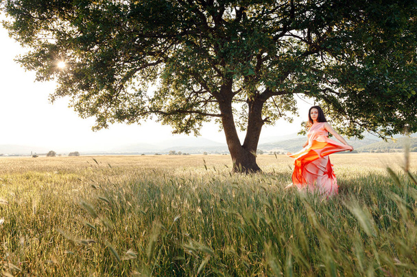 Krásné aktivní zdarma dívka na letním zeleném venkovním pozadí. dívka tančí na poli v krásných růžových šatech u mocného dubu - Fotografie, Obrázek