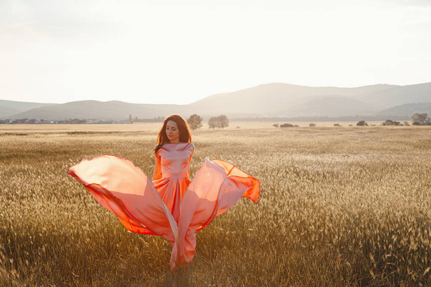 Tyttö tanssii pellolla kauniissa vaaleanpunaisessa mekossa auringonlaskun aikaan. ihanteellinen mainontaan ja valokuva aurinko paistaa kirkas ja mehukas
 - Valokuva, kuva
