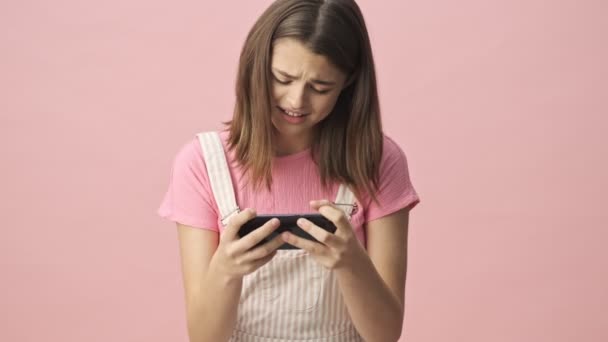 Concentrated pretty brunette woman in overalls playing on smartphone and becoming happy over pink background - Metraje, vídeo