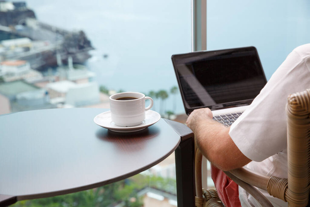 Δουλεύοντας από το σπίτι. Χώρος εργασίας με laptop και φλιτζάνι καφέ στο μπαλκόνι με θέα στον ωκεανό. - Φωτογραφία, εικόνα