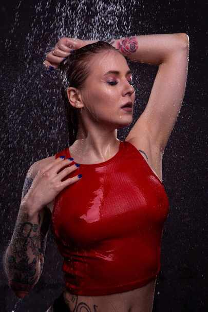 Portrait of a sexy girl with beautiful big Breasts in a wet red t-shirt taking a shower on a dark background with bright drops of water falling. - Photo, Image