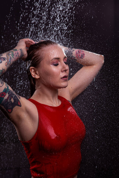 Close-up portret van een sexy meisje met mooie borsten in een nat rood t-shirt op een donkere achtergrond met heldere druppels water die vallen. Reclame, commerciële vormgeving. - Foto, afbeelding