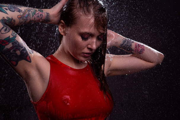 A charming blonde girl with beautiful Breasts in a red wet t-shirt stands under the bright splashes of water straightening her hair. Close-up portrait - Valokuva, kuva