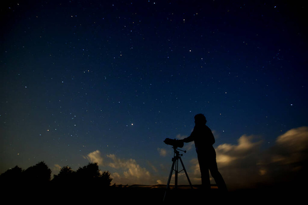 Silhouet van een volwassen man met een telescoop die de sterrenhemel en sterrenstelsels bestudeert. Een man met een astronomisch apparaat staat 's nachts op de achtergrond van een natuurlijk landschap. - Foto, afbeelding