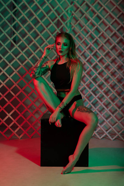 A beautiful tattooed blonde girl is sitting on a cube with her long legs wrapped in her hands. Studio fashion shooting in mixed light - Fotografie, Obrázek