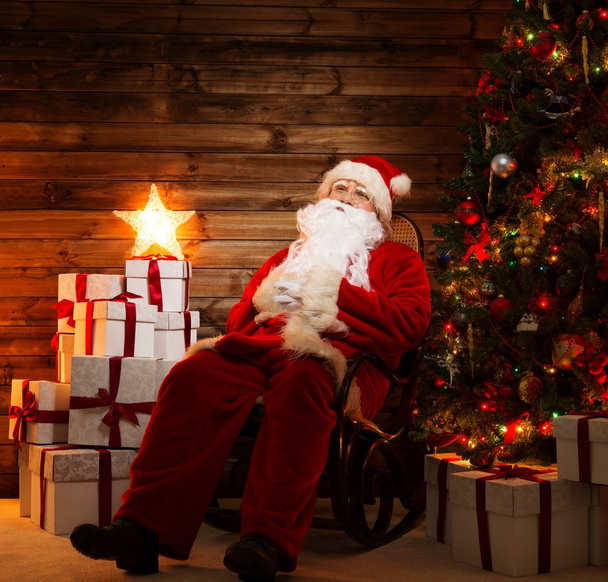 Weihnachtsmann sitzt auf Schaukelstuhl im hölzernen Wohnbereich mit beleuchtetem Stern auf Geschenkschachteln - Foto, Bild