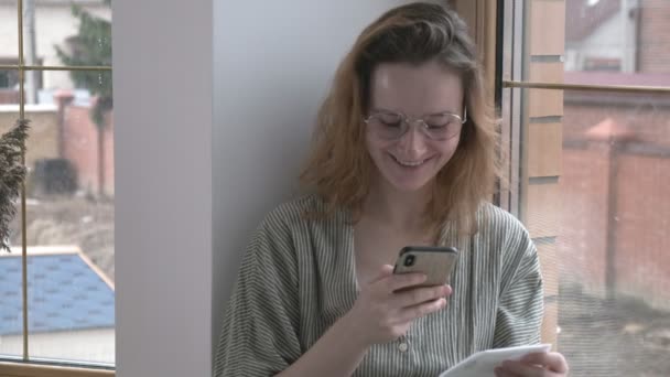 Mädchen mit Brille sitzt auf Fensterbank, chattet per Smartphone. Online-Kommunikation - Filmmaterial, Video