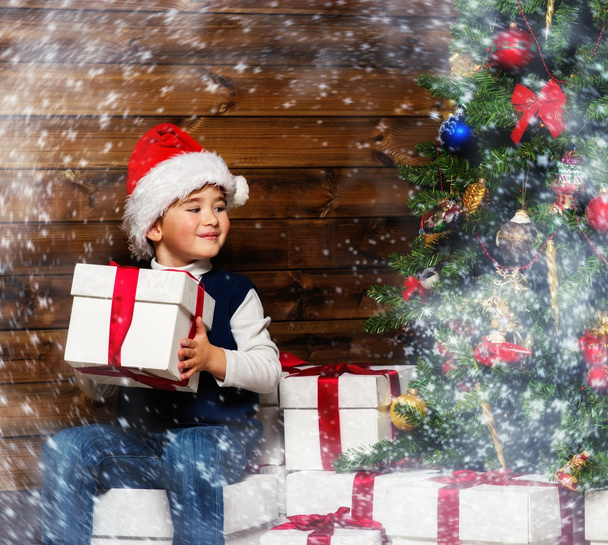 Menino pequeno em chapéu de Santa com caixa de presente sob árvore de natal em interior de casa de madeira
 - Foto, Imagem