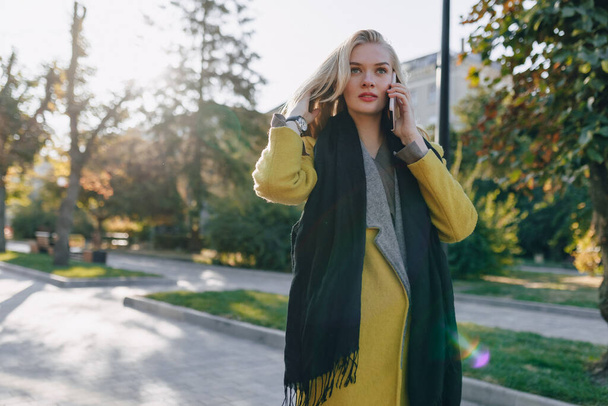 cute emotional attractive blonde woman in coat with smartphone walks the city street. communication during the walk, lifestyle, street in good weather. - Photo, Image