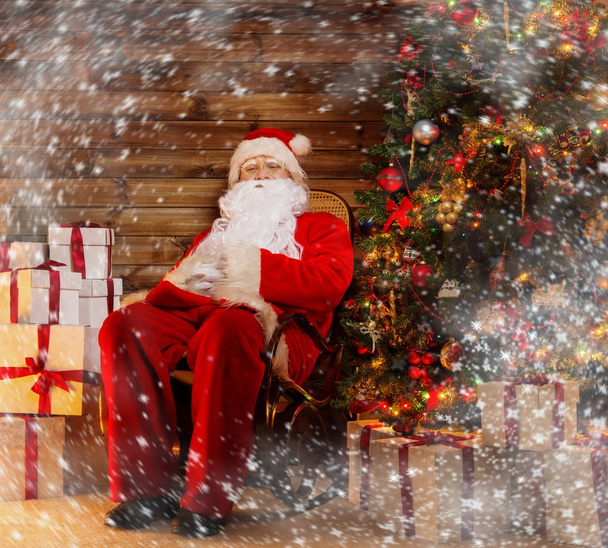 Santa claus zittend op schommelstoel in houten interieur met geschenkdozen om hem heen - Foto, afbeelding