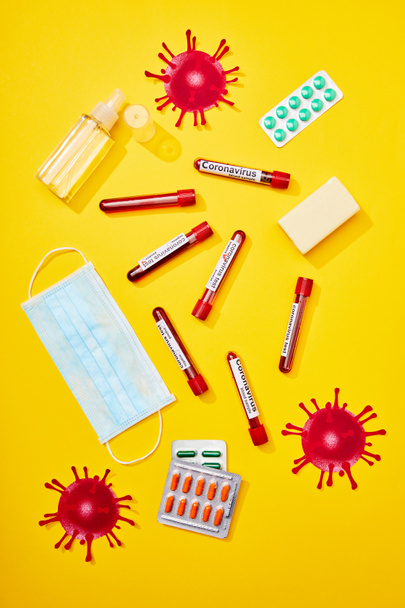 Tıbbi maskenin yanında koronavirüs harfleri olan test tüplerinin üst görüntüsü, haplı su toplamaları, el dezenfektanlı şişe ve sarı üzerine sabun çubuğu.  - Fotoğraf, Görsel