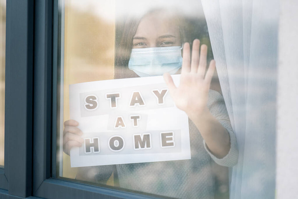 Concept of coronavirus quarantine. Child wearing medical protective face mask during flu virus, looking out of window. COVID-19. Teen girl holds board sign with text Stay at home. View from street. - Foto, immagini
