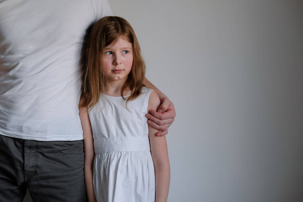 A sad and upset little girl in a white dress stands on a light background next to her dad. angry daughter offended by parent - Fotografie, Obrázek