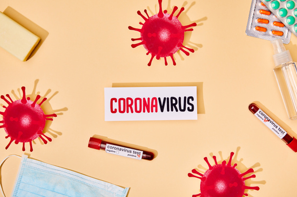 Üzerinde Coronavirus harfleri olan kağıdın üst görüntüsü virüsün yanında, test tüpleri, sabun çubukları, tıbbi maske, haplar ve bej üzerine el dezenfektanı.  - Fotoğraf, Görsel