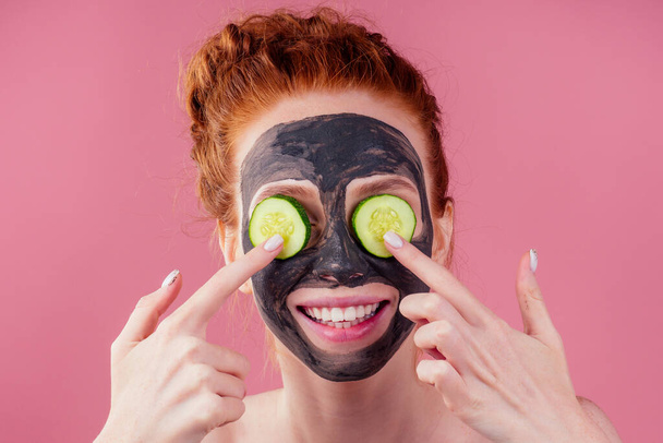 redhead ginger female with cucumber and clay mask on her pretty face, snow white smile in studio pink background - Fotoğraf, Görsel