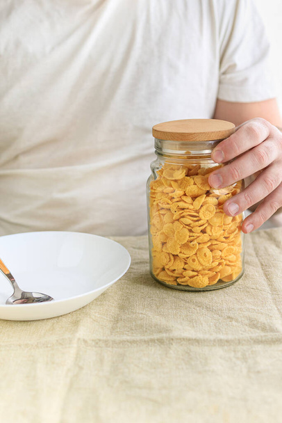 Een gehakte man houdt een pot in zijn handen met cornflakes op een gewoon ruw tafelkleed. Een close-up. Selectieve focus. Concept, eenvoudige snelle Amerikaanse gezonde ontbijt - Foto, afbeelding
