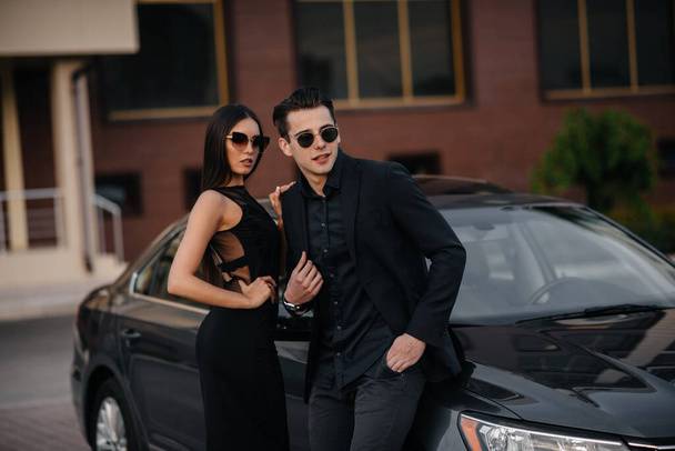 A young stylish couple in black stands near the car at sunset. Fashion and style - Φωτογραφία, εικόνα