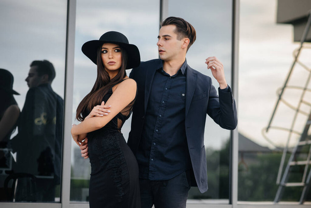 A beautiful, stylish pair of young people in black clothes and glasses stand against the background of an office building in the sunset. Fashion and style - Фото, изображение