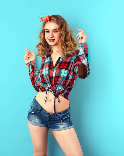 Pin-up ragazza con i capelli biondi ricci con fiocco a scacchi rossi e camicia che indossa pantaloncini jeans. - Foto, immagini
