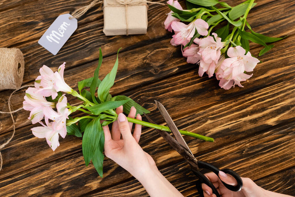 top view of woman holding scissors near pink flowers, gift box and tag with mom lettering, mothers day concept  - Φωτογραφία, εικόνα