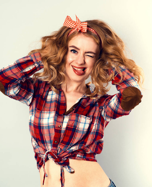 Pin-up Mädchen mit lockigem blondem Haar mit rot karierter Schleife und Hemd in Jeans-Shorts. - Foto, Bild