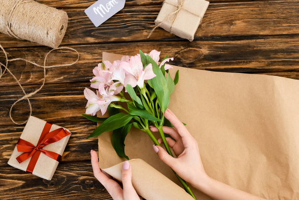 top view of woman holding pink flowers near paper, jute twine rope and gift boxes on wooden surface, mothers day concept  - Photo, Image