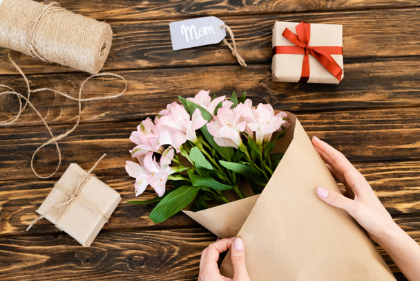 cropped view of woman wrapping pink flowers in paper near gift boxes on wooden surface, mothers day concept  - Foto, Bild