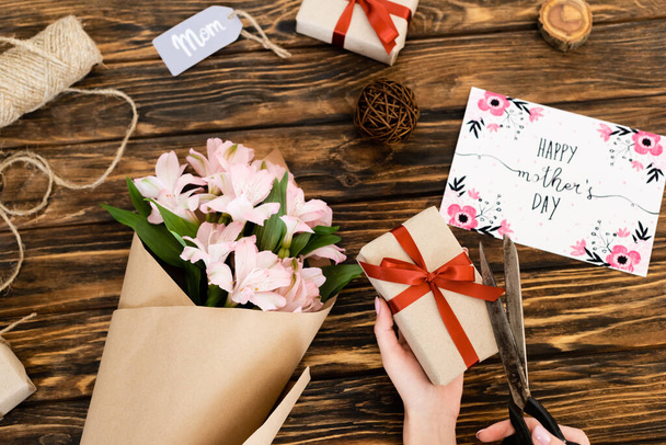 cropped view of woman cutting ribbon on present near pink flowers, greeting card with happy mothers day and tag with mom lettering on wooden surface - Foto, Bild