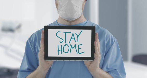 Doctor wearing face mask holds tablet PC with STAY HOME handwritten text. Self-isolation during COVID-19 disease outbreak related shot - Photo, image
