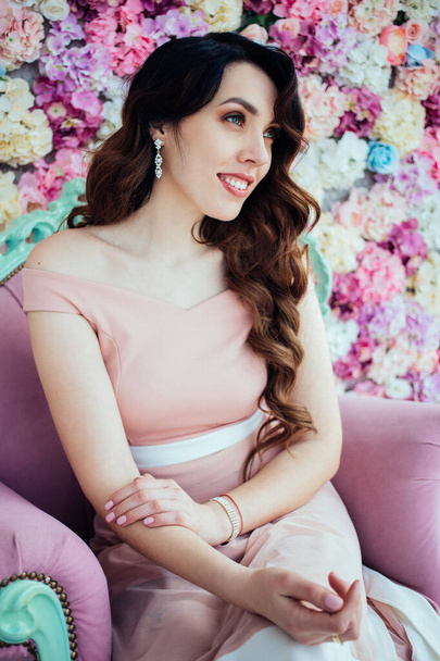 A smiled girl in pink dress with earrings sits on the pink armchair. Flowers background. A girl with beautiful curly dark hair. A cute girl is smiling.  - Photo, Image