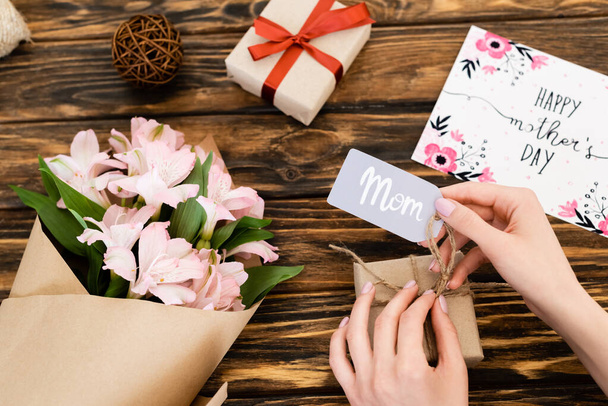 cropped view of woman touching tag with mom lettering on gift box near pink flowers and greeting card with happy mothers day on wooden surface  - Foto, Imagem