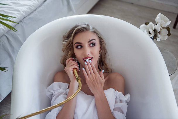 A beautiful young woman in a white wedding dress is lying in the bathroom, talking in the shower like shes on the phone, smiling. The bathroom is in a beautiful interior with green plants - Foto, immagini