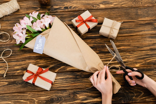 cropped view of woman cutting jute twine rope on pink flowers wrapped in paper near gift boxes and scissors, mothers day concept  - Foto, afbeelding